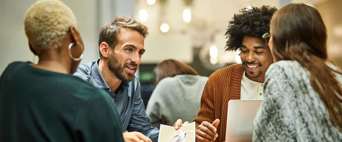 A group of male and female coworkers smiles and talks around a table with a laptop computer.