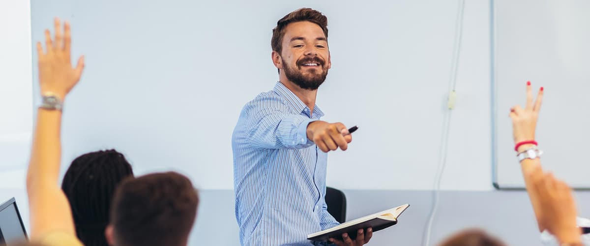 A man is standing in front of a group of students holding a notepad and pointing to a student who is raising their hand.